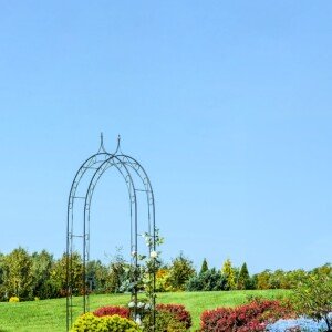 Rosenbogen im Garten vor blauem Himmel mit grüner und bunt blühender Beeteumgebung.